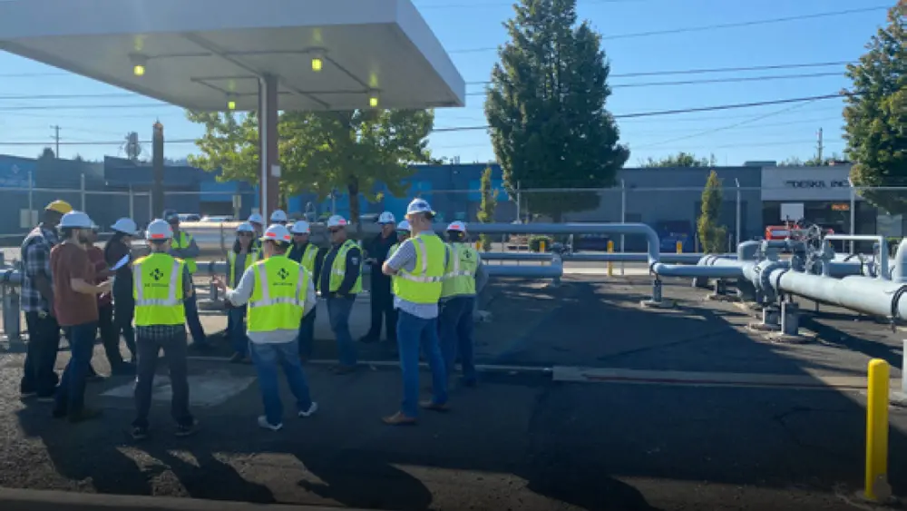 Workers in yellow vests stand at NW Natural facility on pavement in a circle wearing white hard hat discussing on-site hydrogen blending.