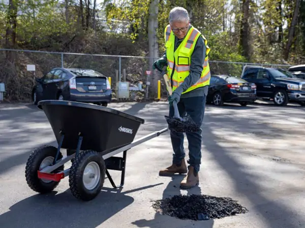 The image shows Bill Gates in a high-visibility yellow safety vest and gloves, using a shovel to place asphalt or similar material into a pothole in a parking lot. He is working with a black wheelbarrow, labeled "Amesbury," filled with the material. Several parked cars and a chain-link fence can be seen in the background, along with trees and vegetation, indicating an outdoor setting. The scene depicts maintenance or repair work to improve the parking lot's surface.