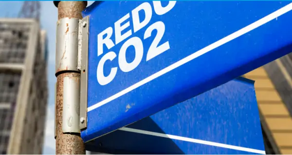 The image shows a close-up of a blue street sign with the white text "REDUCE CO2." The sign is mounted on a rusty metal pole, indicating an effort to promote environmental sustainability and awareness about reducing carbon dioxide emissions. In the blurred background, there are tall buildings, suggesting an urban setting where industrial and urban activities are common. The overall message of the image emphasizes the importance of taking action to lower CO2 levels to combat climate change.