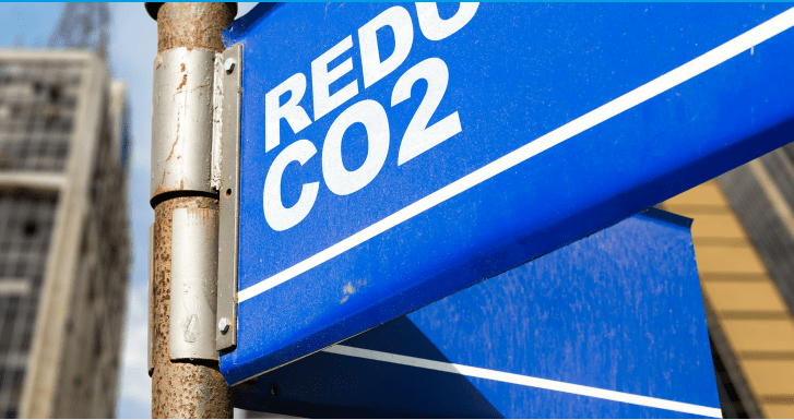 The image shows a close-up of a blue street sign with the white text "REDUCE CO2." The sign is mounted on a rusty metal pole, indicating an effort to promote environmental sustainability and awareness about reducing carbon dioxide emissions. In the blurred background, there are tall buildings, suggesting an urban setting where industrial and urban activities are common. The overall message of the image emphasizes the importance of taking action to lower CO2 levels to combat climate change.