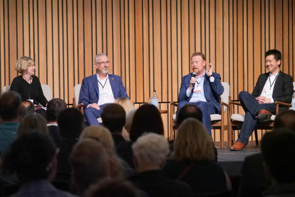 The image shows a panel discussion in progress, with four individuals seated on stage, engaged in conversation. From left to right: A woman with short blonde hair, dressed in black, is smiling and holding a pen and notebook. A man with gray hair and glasses, wearing a blue suit, is looking attentive. A man with reddish-blonde hair and a beard, also in a blue suit, is speaking into a microphone and gesturing with his hand. A man with short black hair, dressed in a black blazer and jeans, is smiling. The audience is visible in the foreground, attentively listening to the discussion. The backdrop consists of vertical wooden slats, adding a professional and warm ambiance to the setting.