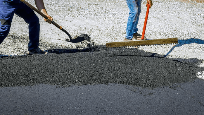 The photo shows two workers spreading and leveling freshly laid asphalt on a road or pathway. One worker is using a shovel to move the asphalt, while the other is using a rake to smooth it out, ensuring an even surface. The background shows a gravel area that has not yet been paved.