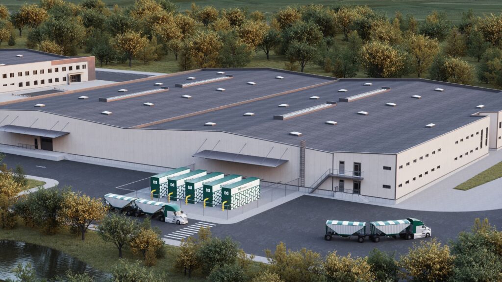 A manufacturing facility surrounded by trees, with several modular green and white distributed natural gas pyrolysis units labeled "Modern Hydrogen" placed outside. The units are connected to a fenced area with visible pipelines. Trucks with green and white accents are parked nearby, including tanker trailers, emphasizing logistics and industrial activity. The setting features a clean, organized layout with paved roads and greenery, blending industrial infrastructure with environmental elements.