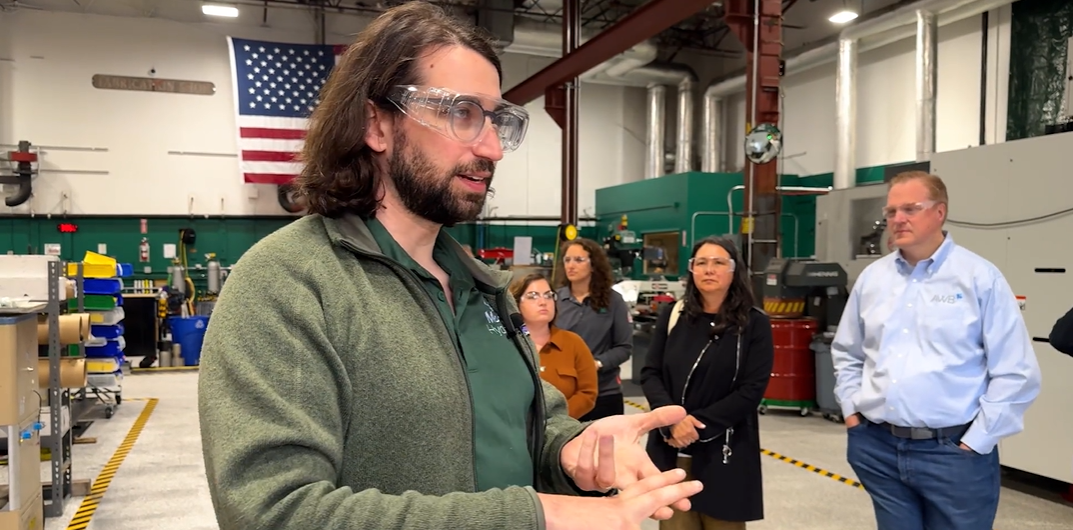 A man with shoulder-length brown hair, a beard, and safety goggles is speaking and gesturing with his hands in an industrial facility. He is wearing a green fleece jacket over a dark green polo shirt. Behind him, a group of four people, including two women in dark clothing, a woman in an orange jacket, and a man in a light blue shirt with "AWB" embroidered on it, are attentively listening. The facility has machinery, tools, and equipment visible, with an American flag hanging on the wall in the background. The floor is clean and organized, with yellow safety markings. CTO Max Mankin is discussing decarbonized natural gas solutions at Modern Hydrogen headquarters.