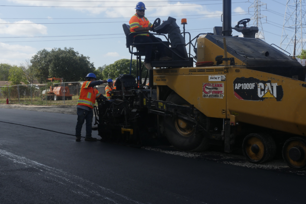 Construction workers in high-visibility vests and blue hard hats operate a large CAT AP1000F asphalt paver, applying Modern Carbon-modified asphalt as part of Clark Construction's sustainability-focused project in San Antonio, Texas. The workers are guiding the material into place, ensuring smooth application. Utility poles, fencing, and construction equipment are visible in the background, emphasizing the infrastructure improvements underway.