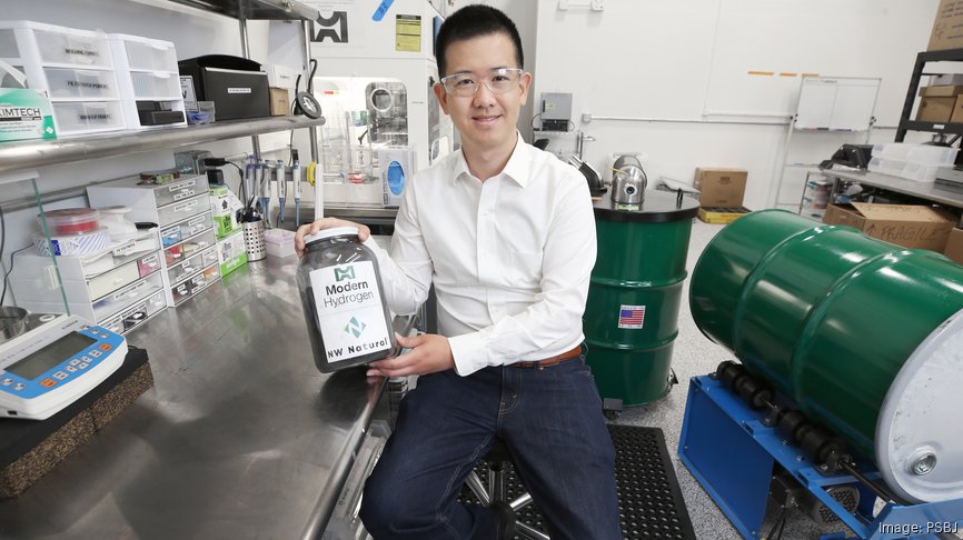 Tony Pan, CEO of Modern Hydrogen, sits in a laboratory wearing safety glasses and a white shirt, holding a clear container labeled "Modern Hydrogen" and "NW Natural" filled with Modern Solid Carbon. Green industrial barrels and lab equipment are visible in the background.