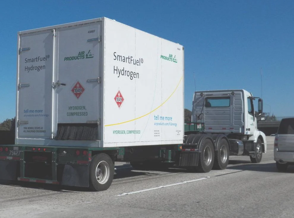 Alt text: A white semi-truck trailer labeled "SmartFuel® Hydrogen" and "Air Products" transports compressed hydrogen on a highway. The trailer displays hazard placards marked "1049" indicating compressed hydrogen gas, and includes promotional text reading "tell me more airproducts.com/h2energy." The truck is driving under clear blue skies.